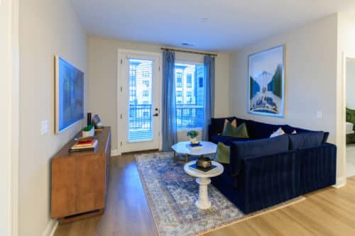 A modern living room with a navy sectional sofa, white coffee table, wooden sideboard, large rug, and framed artwork, featuring glass doors opening to a balcony.
