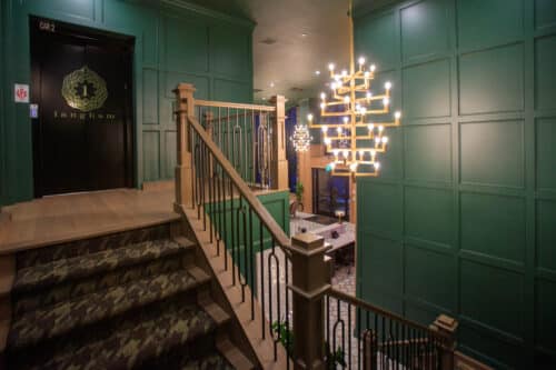 A staircase with patterned carpet leads to a landing with green paneled walls, a black elevator door marked "Langham," and modern chandeliers overhead.