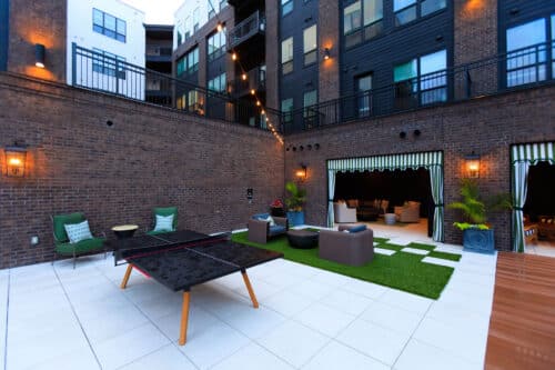 Outdoor patio area with a ping pong table, lounge chairs, string lights, artificial grass, and seating spaces by a brick apartment building.