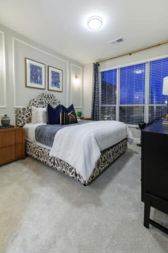 Modern bedroom with a patterned bed, white bedding, decorative pillows, nightstands, artwork on the wall, carpet flooring, and large windows with blinds and blue curtains.