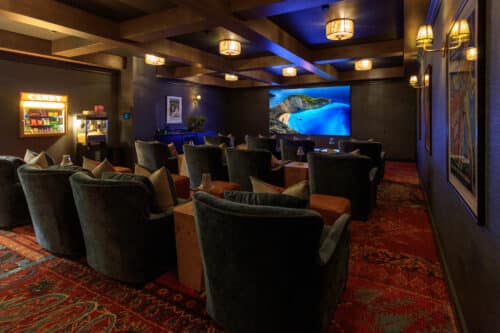 A home theater room with plush chairs facing a large screen displaying a nature scene, dim lighting, and a candy counter on the left side.