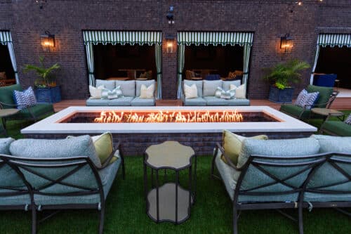 Outdoor patio with cushioned seating around a rectangular fire pit, set on grass, with striped awnings and potted plants in the background.