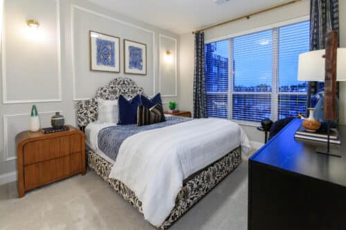 A neatly made bed with patterned headboard, blue and white bedding, two nightstands, a dresser, and large window with blinds in a modern bedroom.