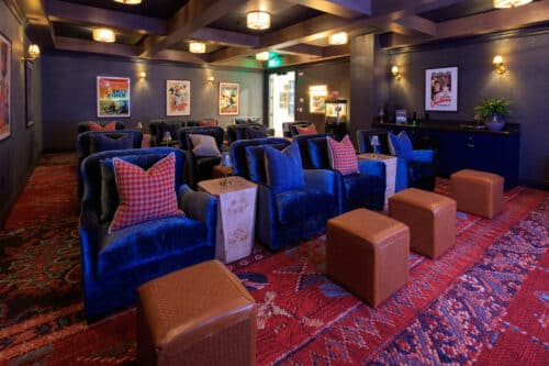 A private home theater room with blue velvet armchairs, brown ottomans, framed movie posters on the walls, and patterned red carpet.