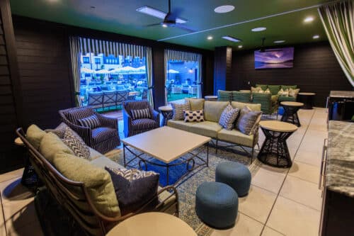 A modern lounge area with cushioned seating, tables, and poufs, opens to an outdoor patio with pool. The room has dark walls, green ceiling, and a wall-mounted TV.