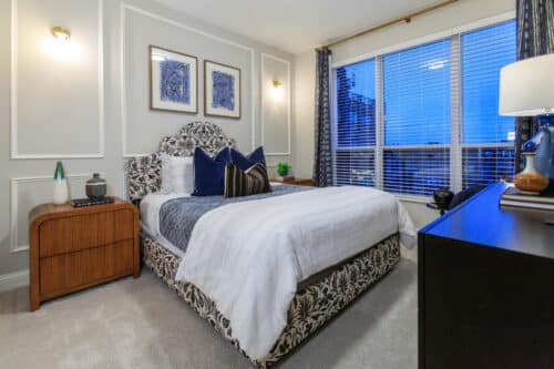 Modern bedroom with a patterned bed, white bedding, wooden nightstands, large window with blinds, and blue-themed decor.