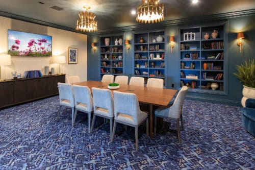 A conference room with a wooden table, ten upholstered chairs, blue carpet, built-in bookshelves, wall sconces, and a large photo of flowers on the wall.
