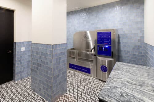 A modern pet washing station with tiled walls and floors, featuring a stainless steel washing unit and touchscreen controls in a well-lit room.