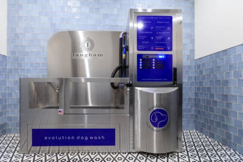 Stainless steel self-service dog wash station with blue instructions and labeled compartments, set against light blue tile walls and patterned floor.