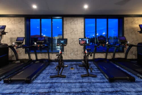 A modern gym room with two stationary bikes and three treadmills facing large windows, each machine equipped with a digital screen.