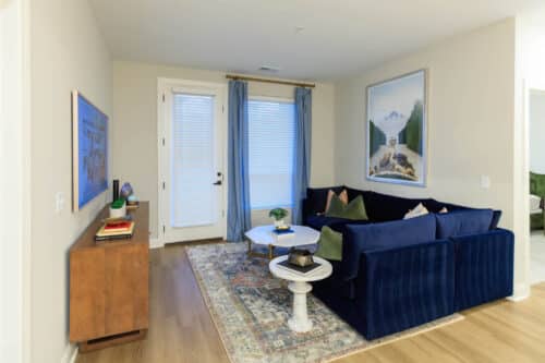 A small living room with a blue velvet sofa, marble coffee table, patterned rug, wooden cabinet, and framed landscape art on cream-colored walls.