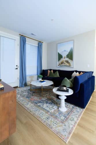 Living room with a blue sectional sofa, patterned rug, white marble coffee table, round side table, blue curtains, and a landscape painting on the wall.