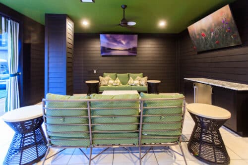 Modern indoor lounge with green cushioned seating, two circular side tables, wall art, ceiling fan, and a countertop under bright ceiling lights.