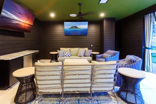 Modern indoor lounge with cushioned seating, striped armchairs, side tables, two wall-mounted TVs, and dark paneled walls. Doorway to the right opens to an outdoor area.
