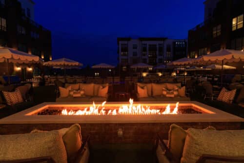 Outdoor patio with cushioned seating around a rectangular fire pit at night, flanked by umbrellas and apartment buildings in the background.