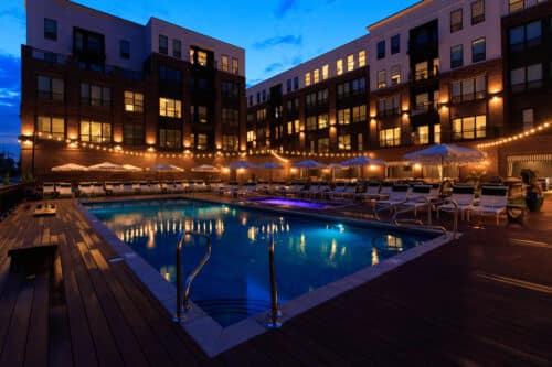 Outdoor swimming pool surrounded by lounge chairs, umbrellas, and string lights, set within a modern apartment complex at dusk.
