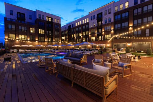 Outdoor pool area at an apartment complex at dusk, featuring lounge seating, string lights, and sun umbrellas with surrounding multi-story buildings.