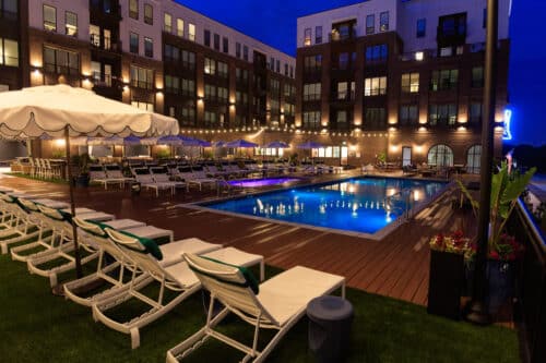 Outdoor swimming pool surrounded by lounge chairs and umbrellas, set against a multi-story apartment building at night with string lights illuminating the area.