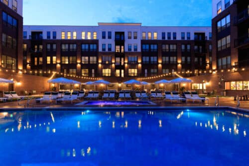 Outdoor swimming pool lit at dusk with lounge chairs, umbrellas, and string lights in front of a modern apartment building.