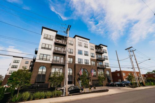 Modern four-story apartment building with balconies, parked cars, and American flags at the entrance, situated on a street with power lines under a partly cloudy sky.