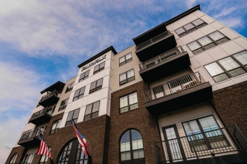 A modern four-story apartment building with balconies, brick and white exterior, and two flags displayed at the entrance under a partly cloudy sky.