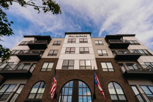 Low-angle view of a modern four-story apartment building labeled "Fairfax," with two flags, balconies, and a partly cloudy sky.