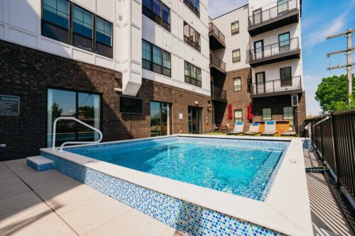 Outdoor rectangular pool beside a modern apartment building, with lounge chairs, patio umbrellas, and a pool rules sign visible nearby.