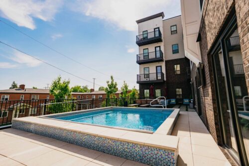 Rectangular above-ground pool on a sunny patio, adjacent to a modern apartment building with balconies and outdoor seating.