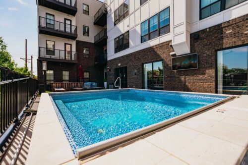 Outdoor rectangular swimming pool next to a modern apartment building with large windows, balconies, and a black metal fence surrounding the pool area.