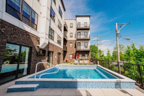 Rectangular outdoor swimming pool beside a modern apartment building with balconies, glass doors, and clear blue sky overhead.