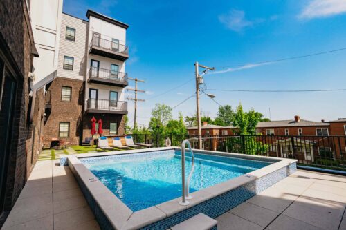 Outdoor apartment pool area with lounge chairs, sun umbrellas, and modern building in the background on a clear, sunny day.