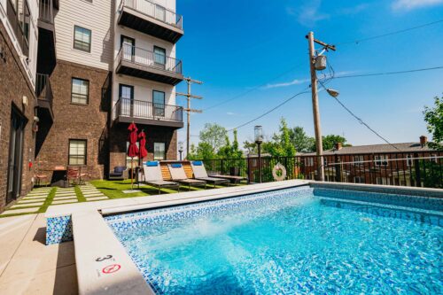 Outdoor pool beside a modern apartment building, with lounge chairs, umbrellas, and clear blue sky in the background.