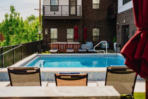 Outdoor swimming pool with poolside lounge chairs, tables, and umbrellas, adjacent to a brick apartment building under daylight.