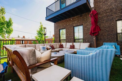 Outdoor patio with cushioned seating, blue chairs, a wooden bench, red umbrella, and brick building in the background on a sunny day.