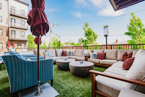Outdoor lounge area with a large sectional sofa, round coffee tables, blue armchair, and closed umbrella on artificial grass, surrounded by a fence and trees, with apartment buildings in the background.