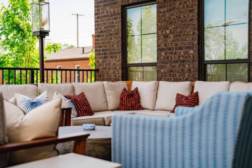 Outdoor patio with beige sectional sofa, red-striped pillows, a round table, blue armchair, and a brick wall with windows in the background.