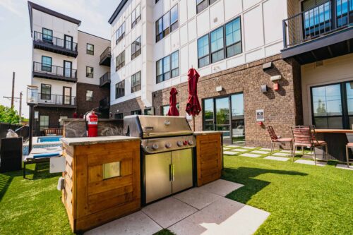 Outdoor grilling area with a stainless steel barbecue, wooden counter, fire extinguisher, and patio seating, set on artificial grass near a modern apartment building.